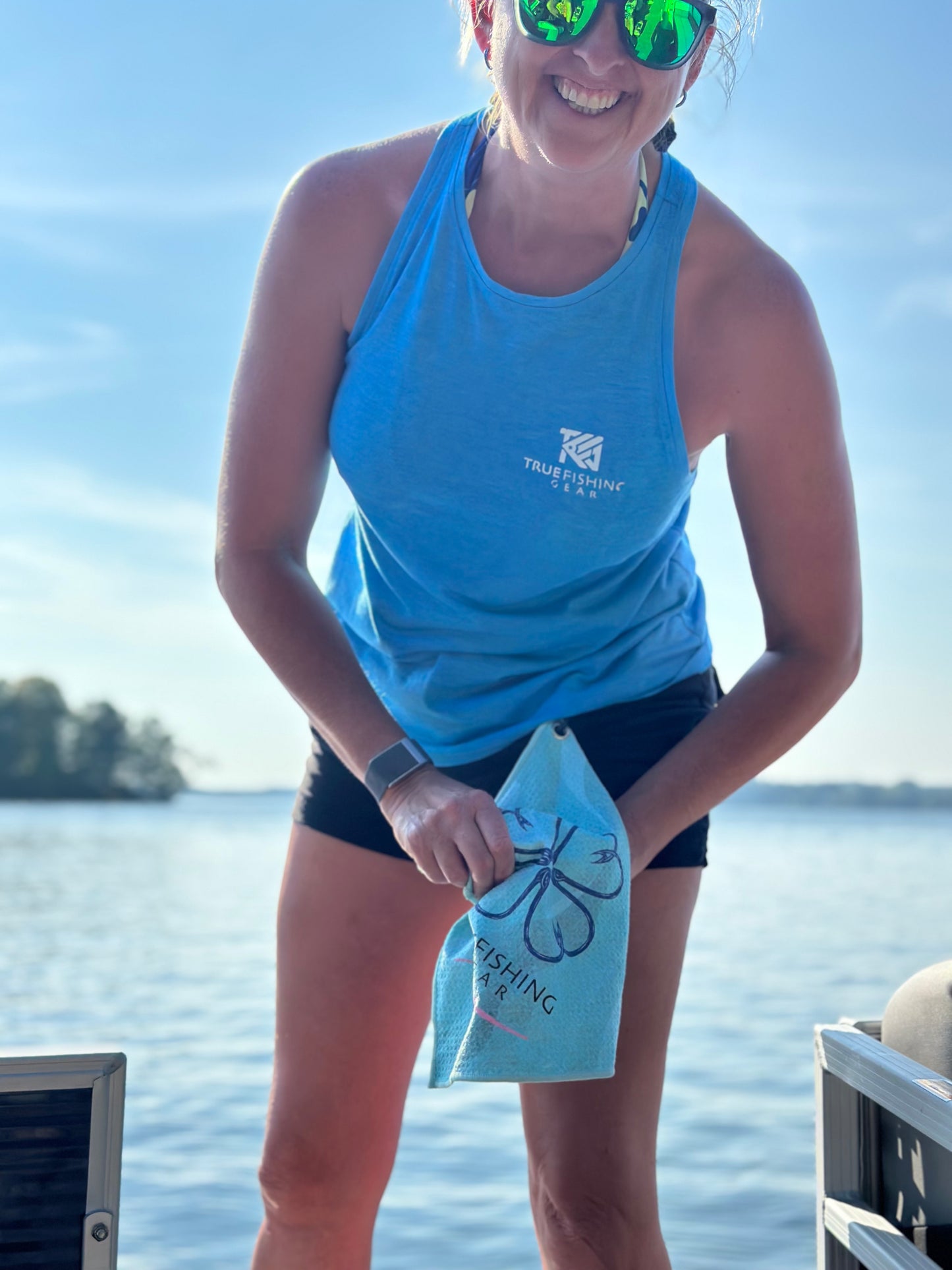 Woman in blue tank top and black shorts wiping hands with a fishing towel by a lake