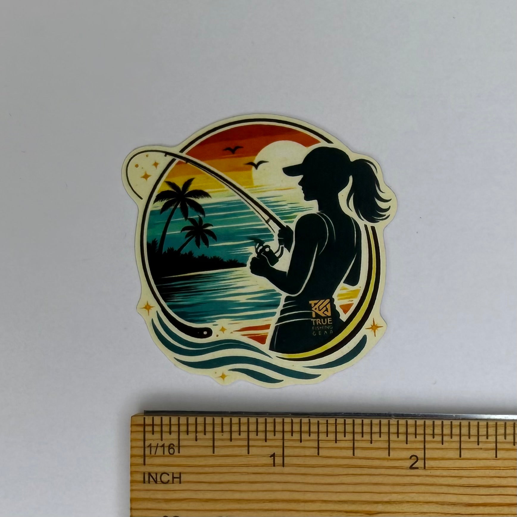 Sticker of a silhouette of a woman fishing with a scenic background, next to a ruler for scale.